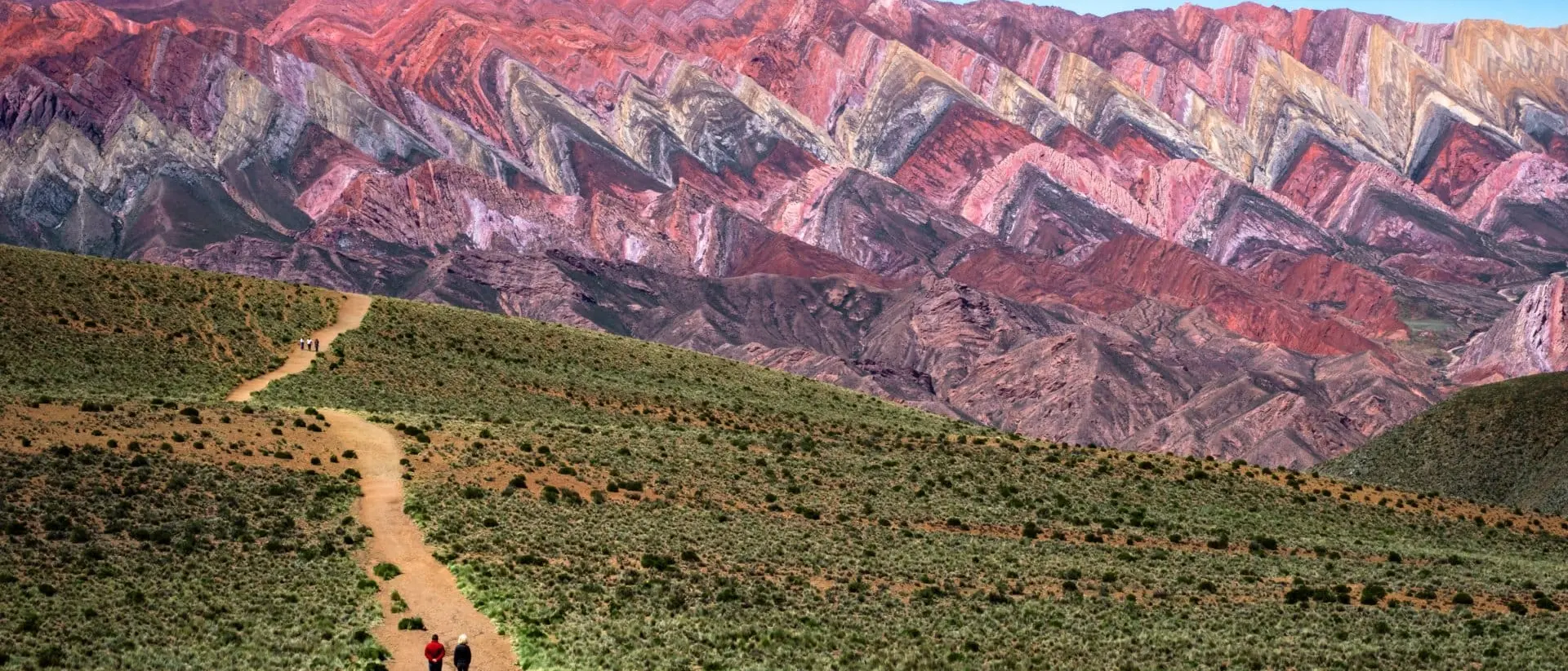 Two hikers walk along a dirt path with colorful striped mountains in the background under a clear blue sky.