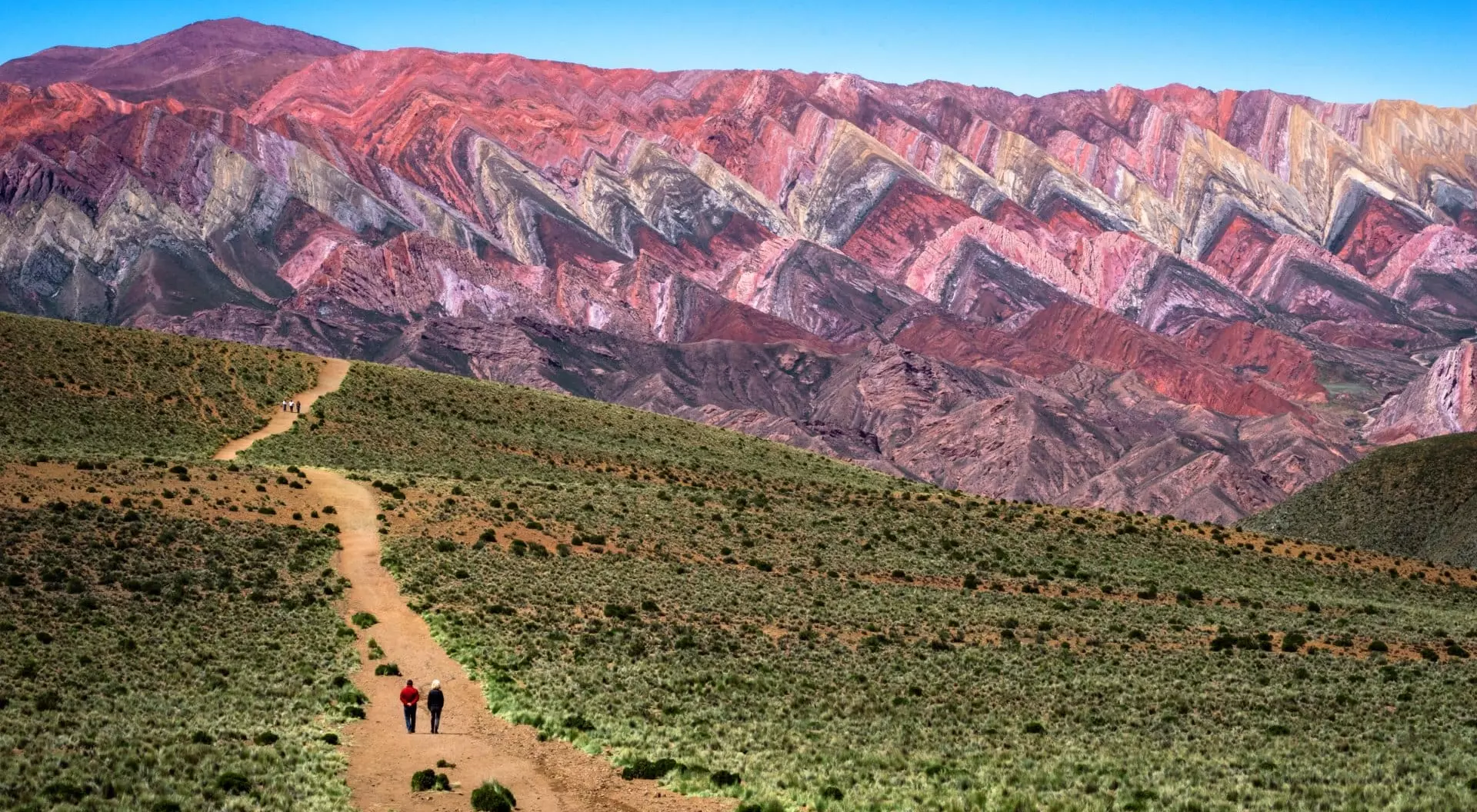 Two hikers walk along a dirt path with colorful striped mountains in the background under a clear blue sky.