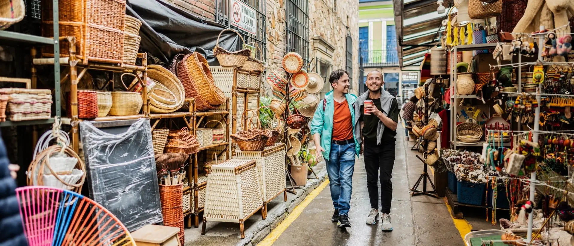 Two men stroll through a vibrant market alley filled with colorful handmade crafts and woven baskets.