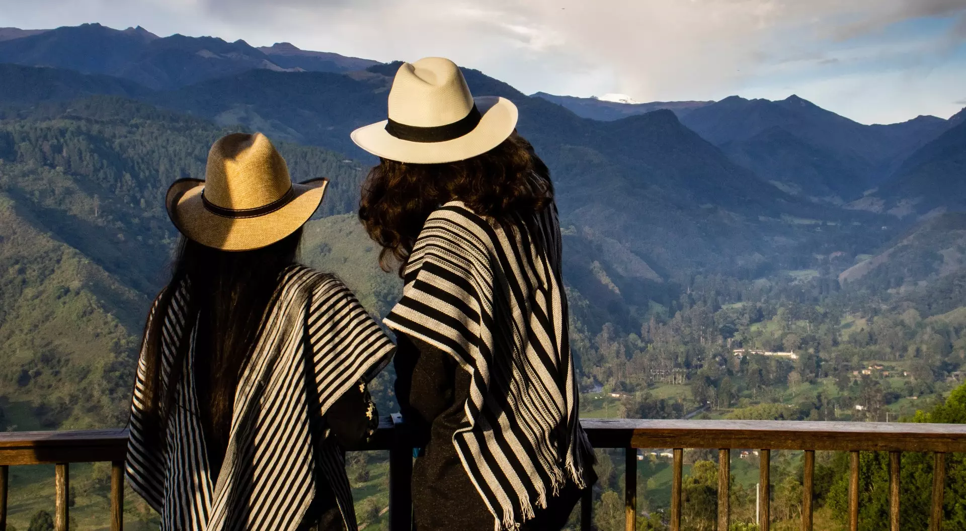 Two people in traditional ponchos and hats stand overlooking a mountainous landscape.