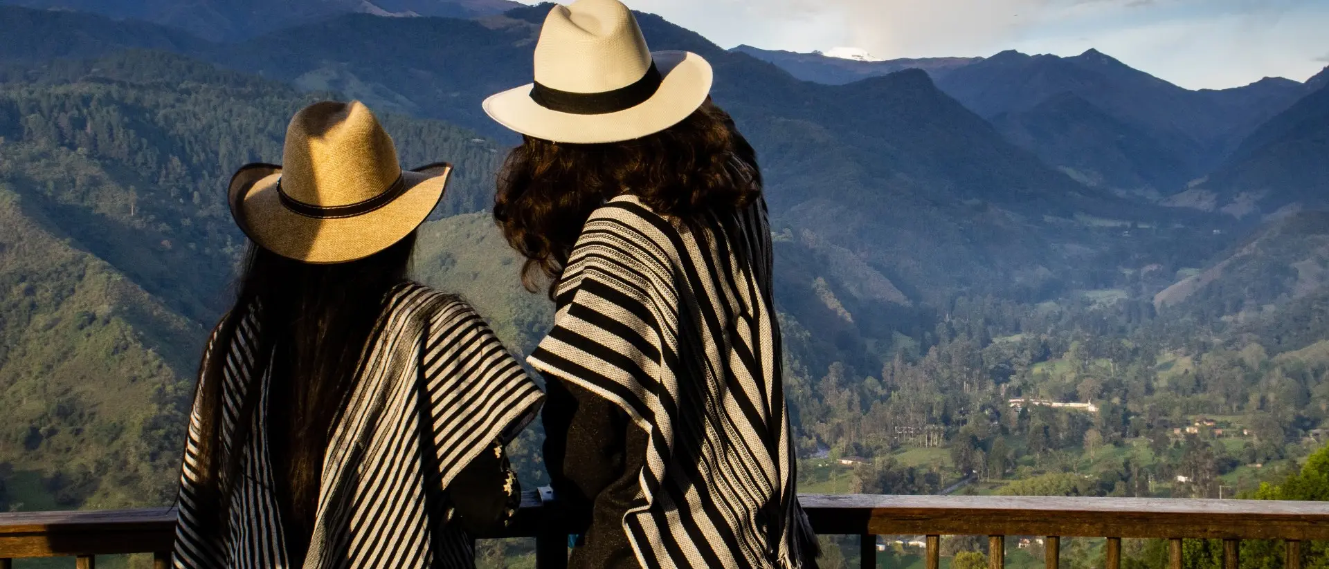 Two people in traditional ponchos and hats stand overlooking a mountainous landscape.