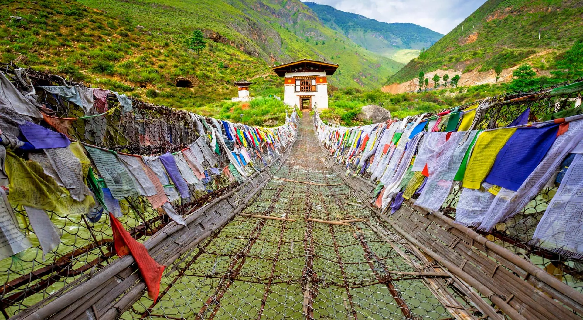 Tamchog Lhakhang View straight on of Iron Chain Bridge of Tamchog Lhakhang Monastery with prayer flags lining the bridge, mountains and cloudy sky in the background