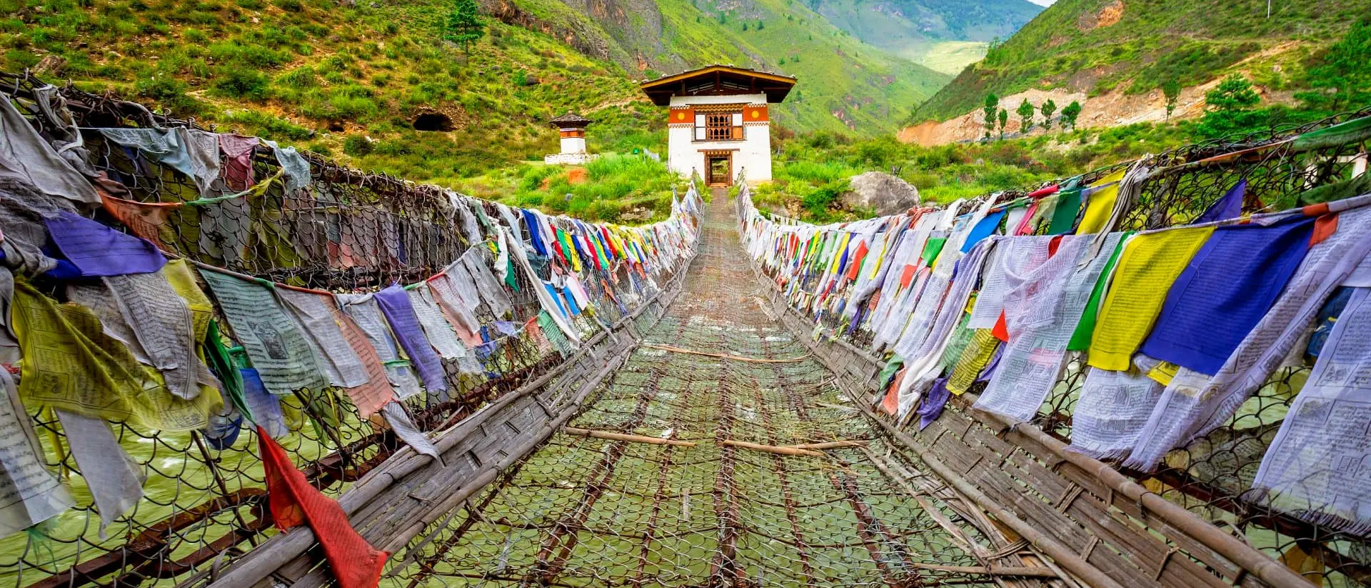 Tamchog Lhakhang View straight on of Iron Chain Bridge of Tamchog Lhakhang Monastery with prayer flags lining the bridge, mountains and cloudy sky in the background
