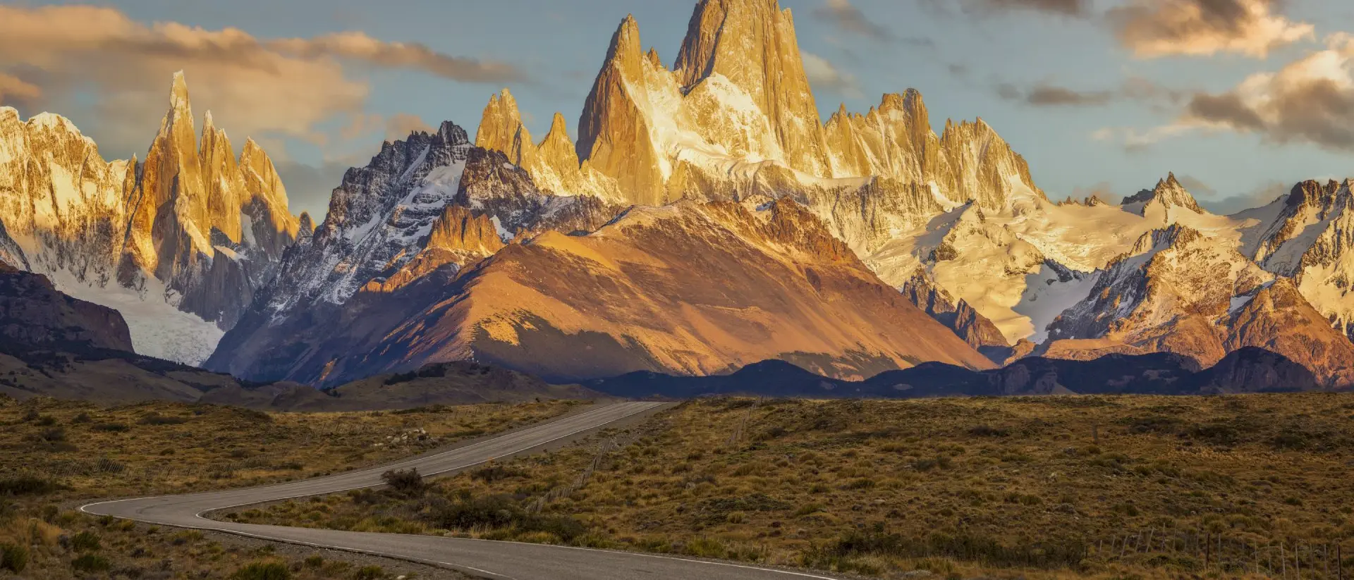 Winding road leads toward dramatic, snow-capped mountains at sunset, with warm, glowing light illuminating the peaks.