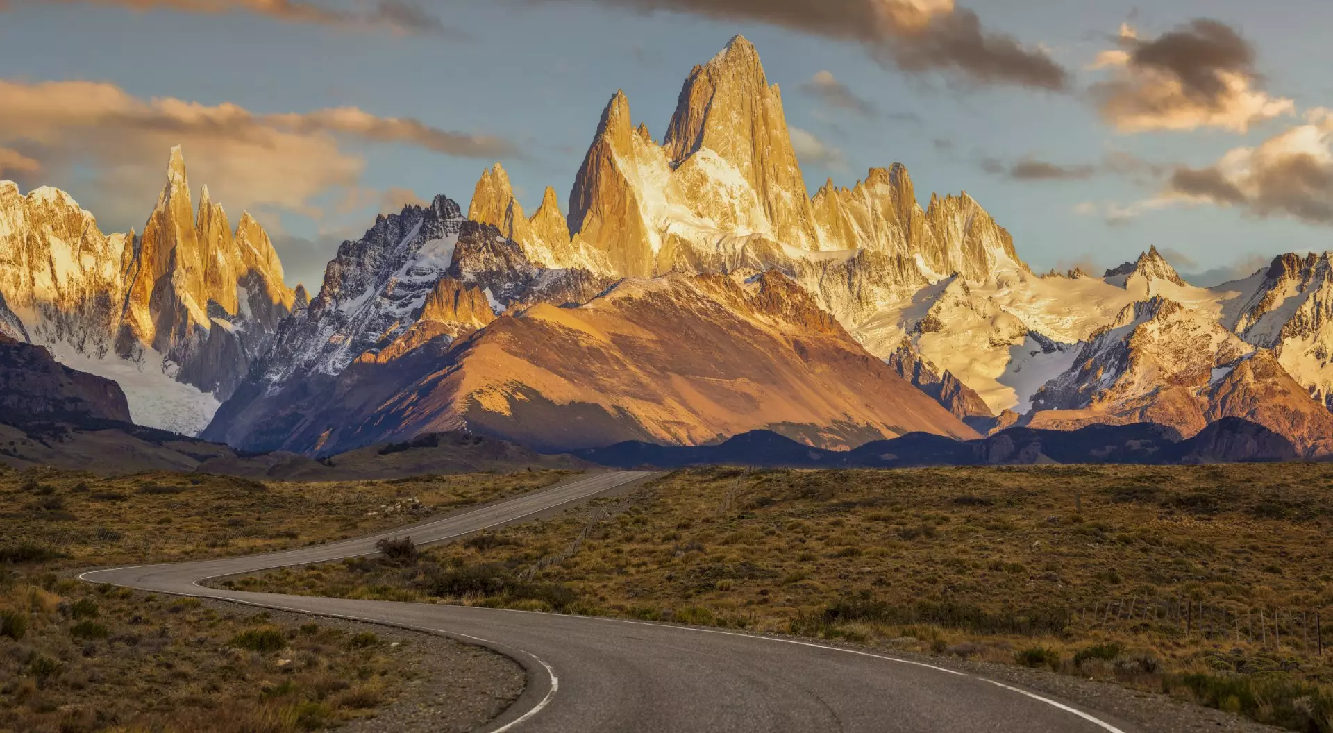 Argentina Winding road leads toward dramatic, snow-capped mountains at sunset, with warm, glowing light illuminating the peaks.