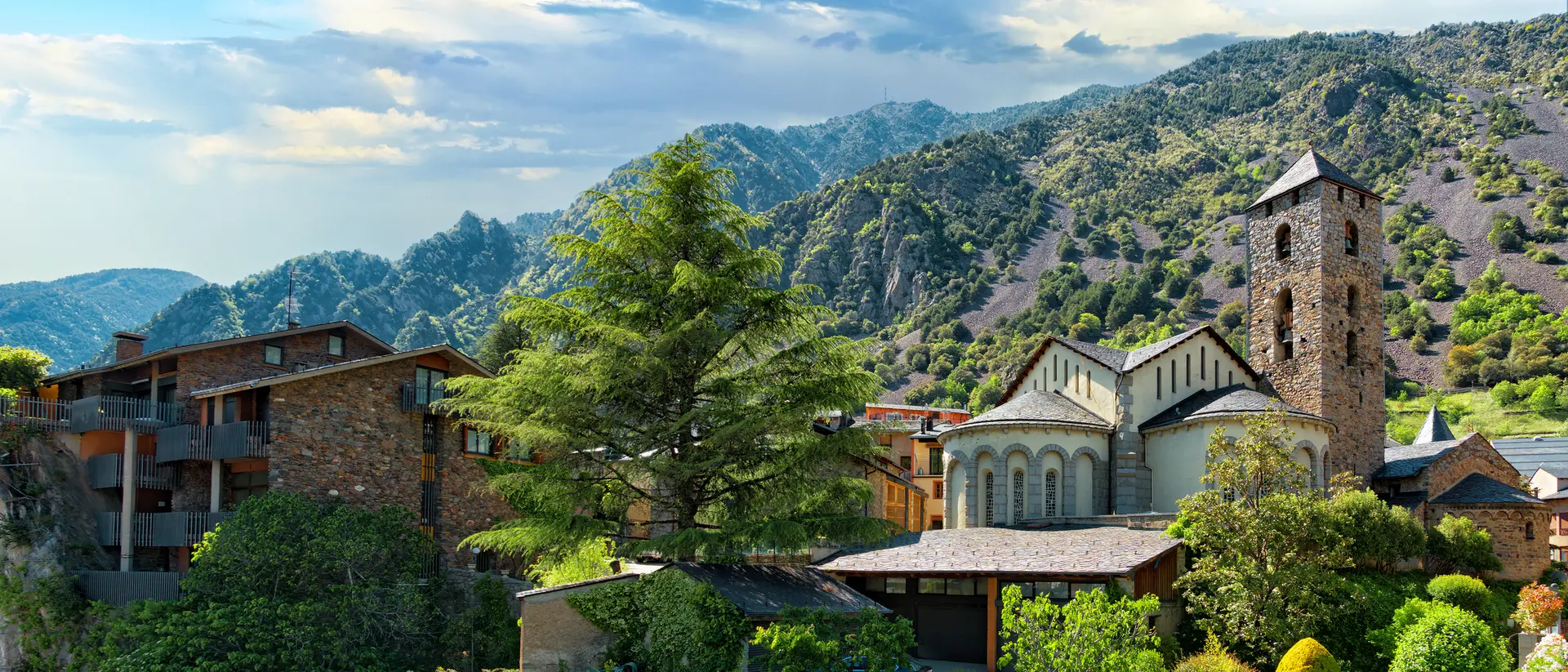 Bright and sunny day over looking lush tree-tops, a mountain range, and a gothic-style building in La Vella, Andorra.