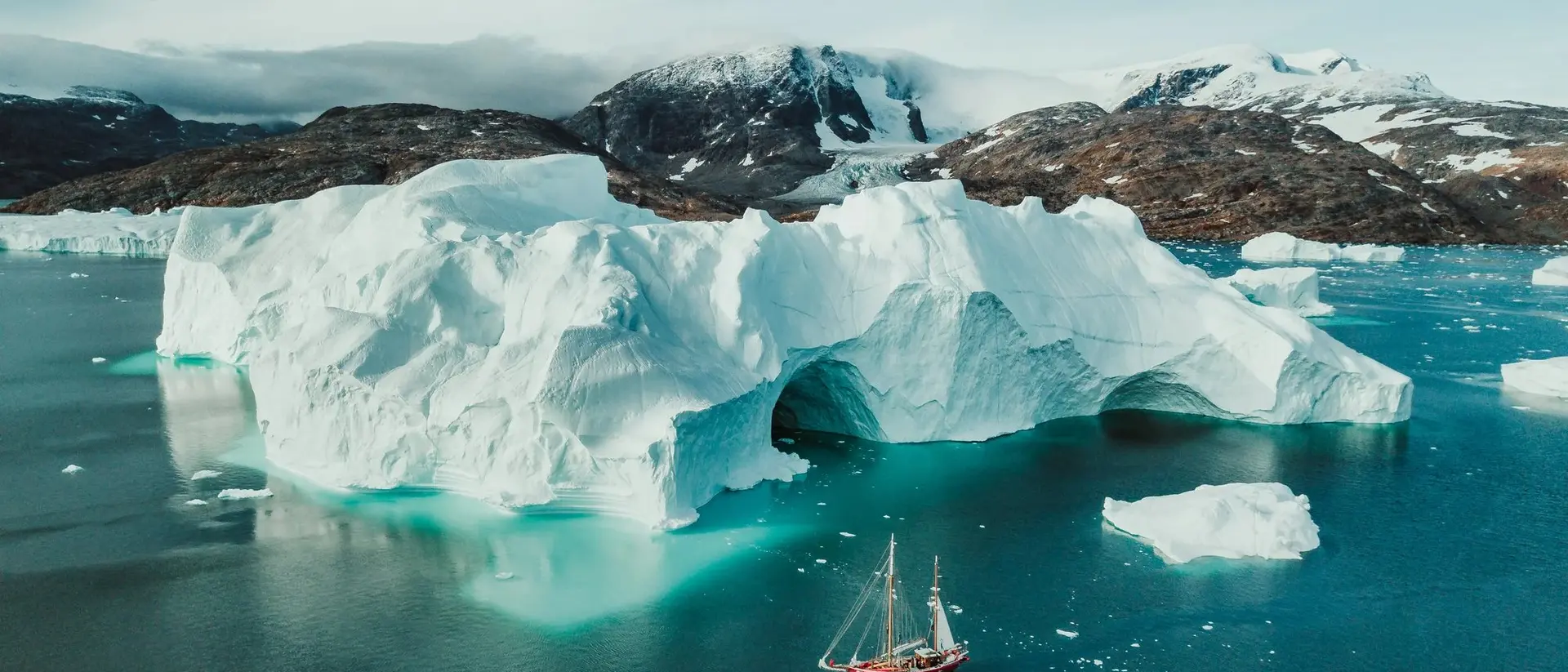 An aerial view of a sailboat passing alongside icebergs in the cold Arctic waters of Greenland.