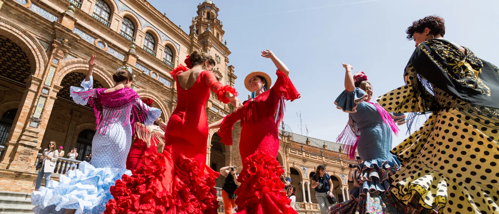 A group of women in colorful flamenco dresses dance in a plaza with historic architecture in the background.