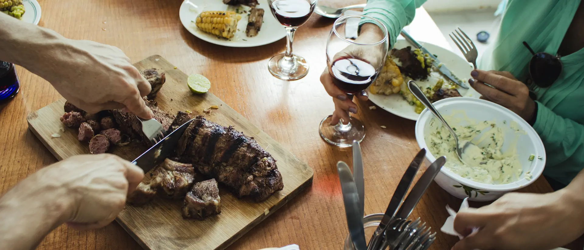 A wood table laden with plates of food, glasses of wine, and hands sharing a meal together.