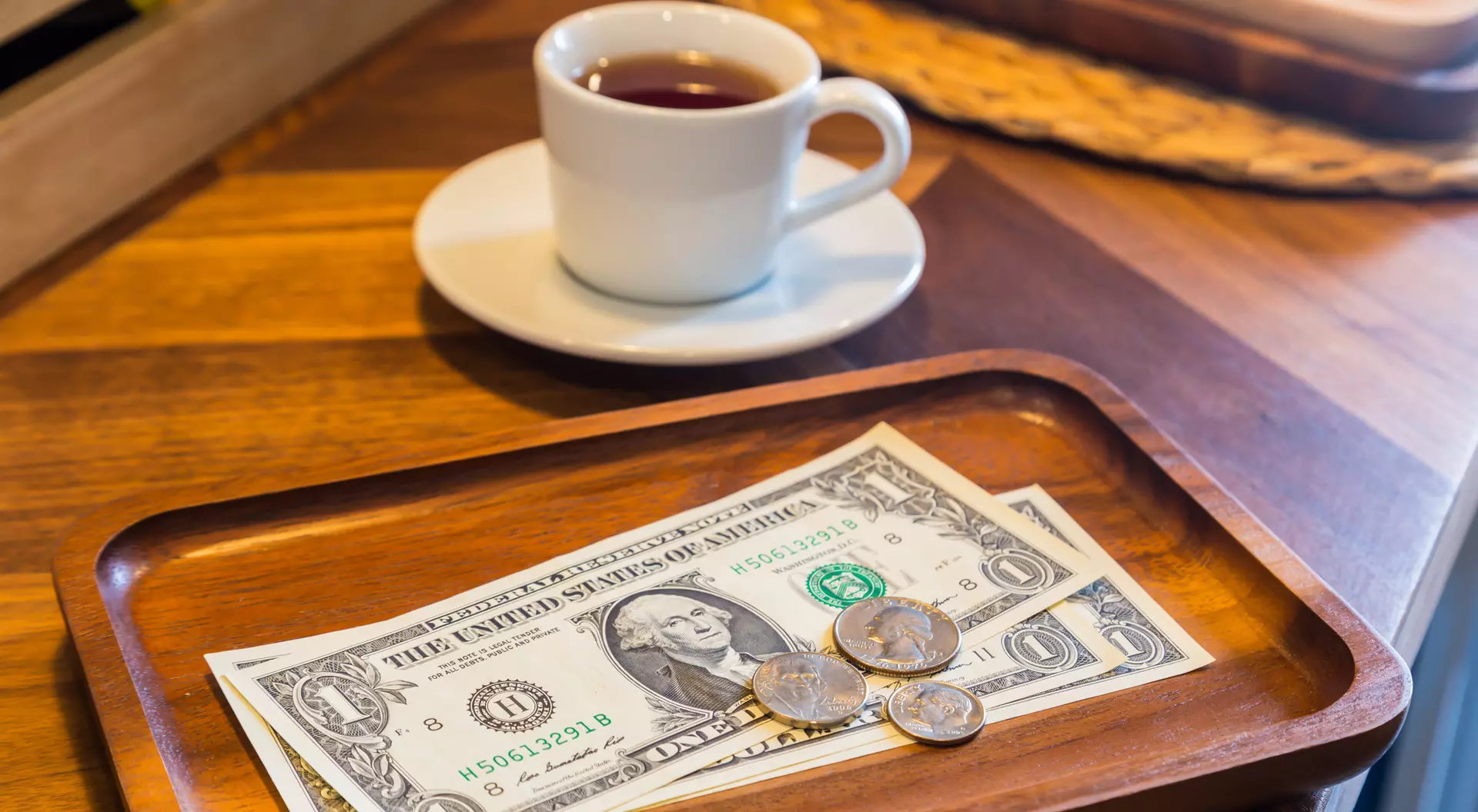 A wooden tray holds a dollar bill and coins, beside a white cup of tea on a wooden table.