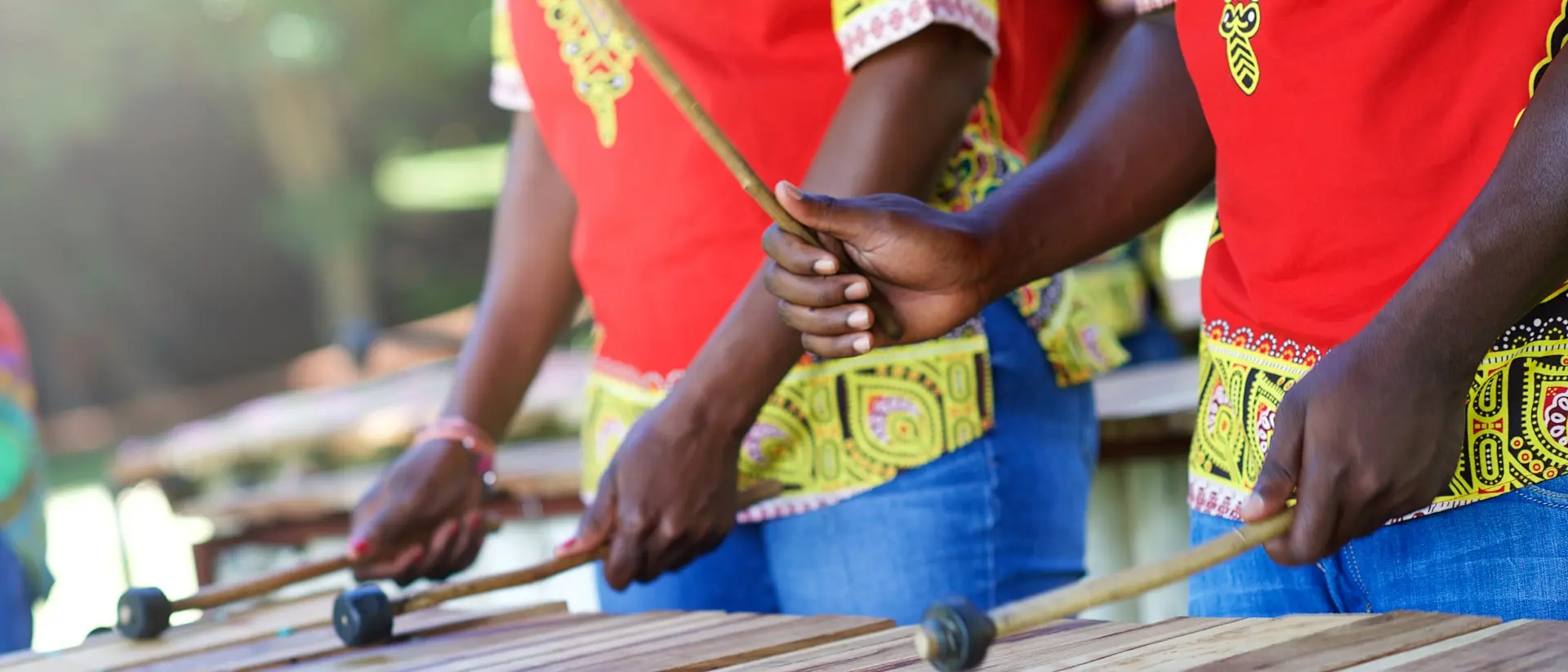 Three musicians in colorful shirts play a wooden xylophone outdoors, holding mallets and striking the bars.
