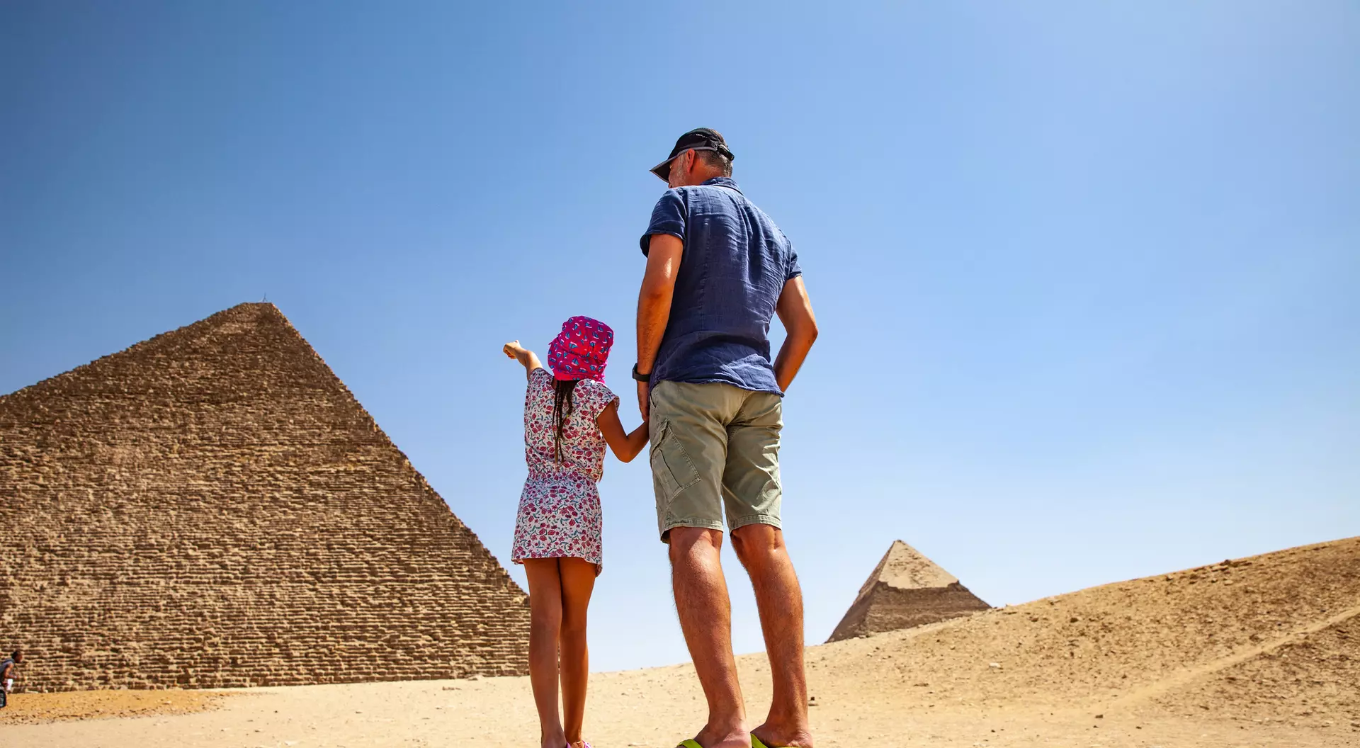 A man and a girl stand together, admiring the Great Pyramids under a clear blue sky.