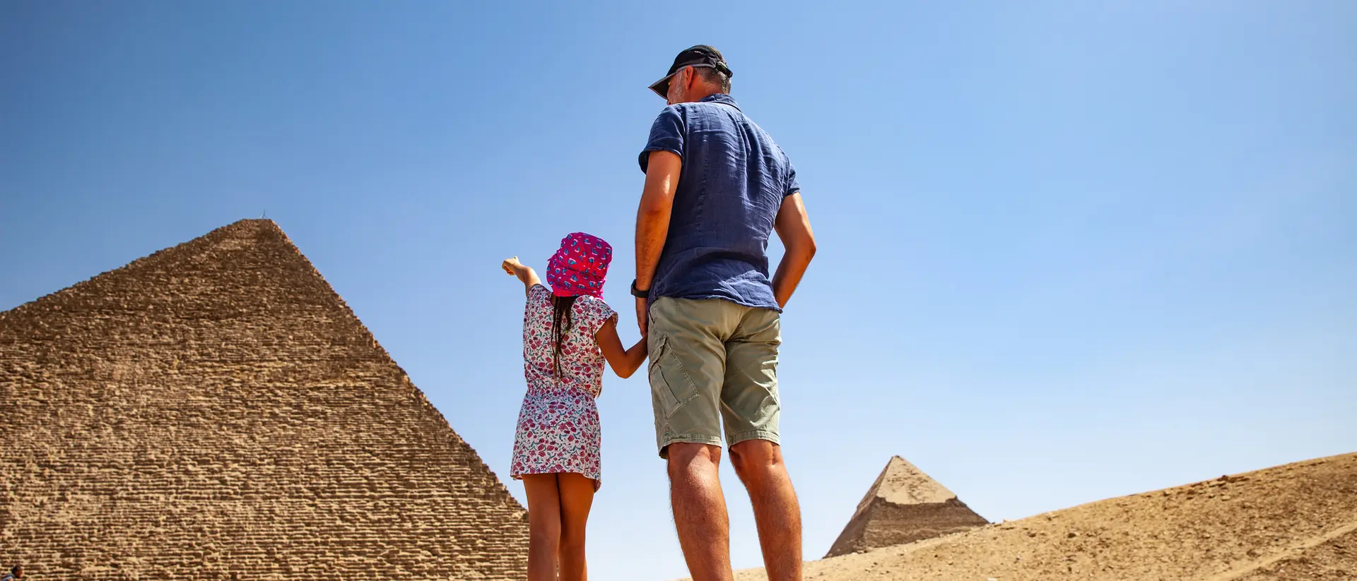 A man and a girl stand together, admiring the Great Pyramids under a clear blue sky.