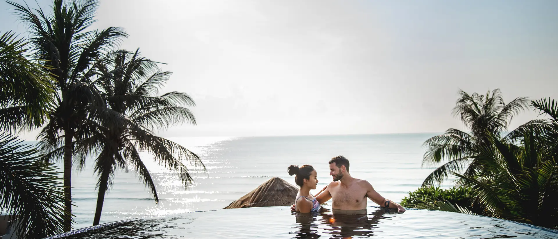 A couple relaxes in an infinity pool overlooking the ocean, surrounded by palm trees under a sunny sky.