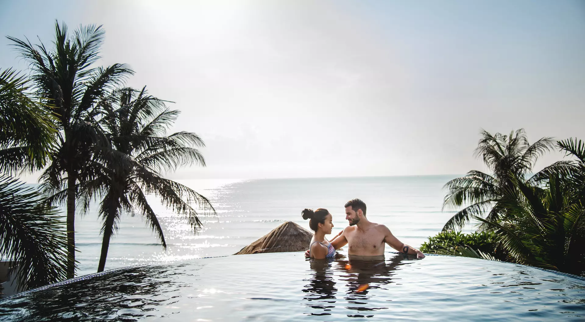 A couple relaxes in an infinity pool overlooking the ocean, surrounded by palm trees under a sunny sky.
