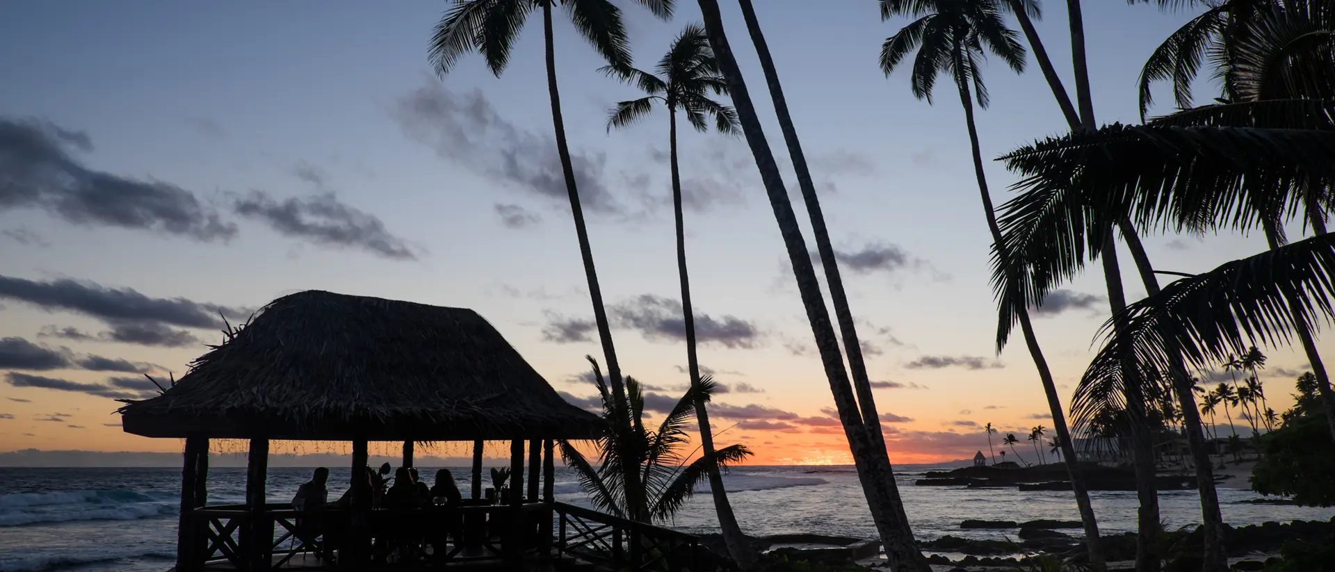 Silhouette of a beach pavilion at sunset, surrounded by palm trees and ocean waves.