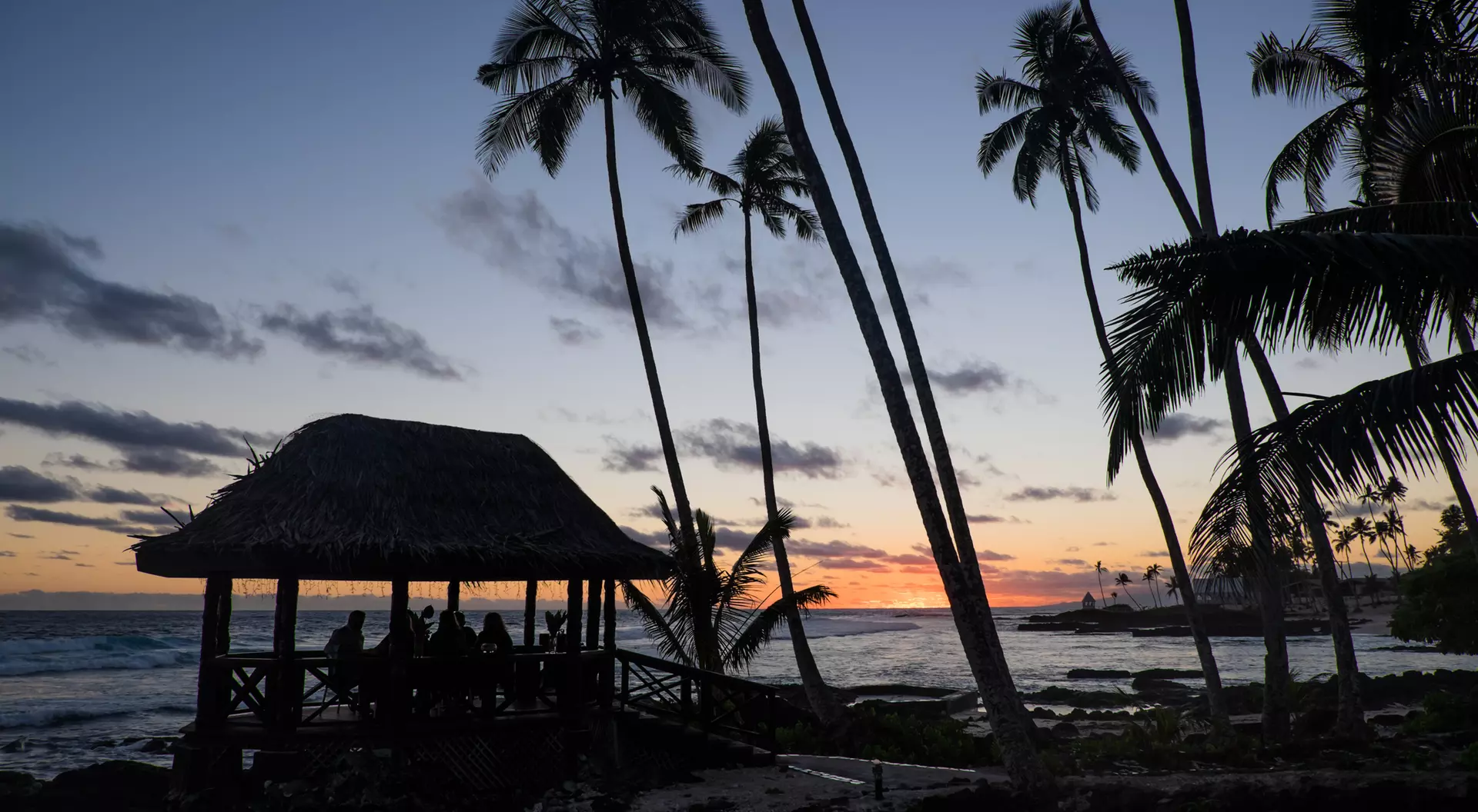 Silhouette of a beach pavilion at sunset, surrounded by palm trees and ocean waves.