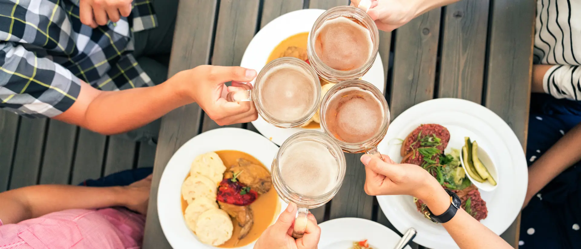 Friends toasting with beer mugs over a table of diverse dishes, enjoying a meal together.