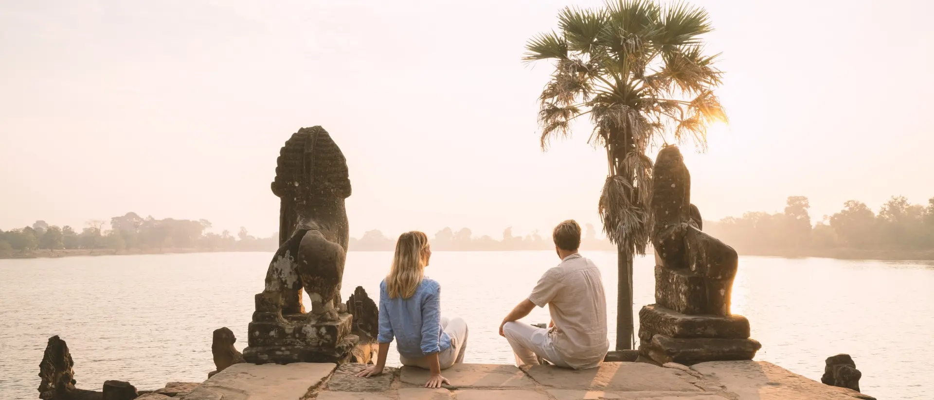 A couple sits on a stone platform, overlooking a calm river at sunrise, flanked by lion statues and a palm tree.