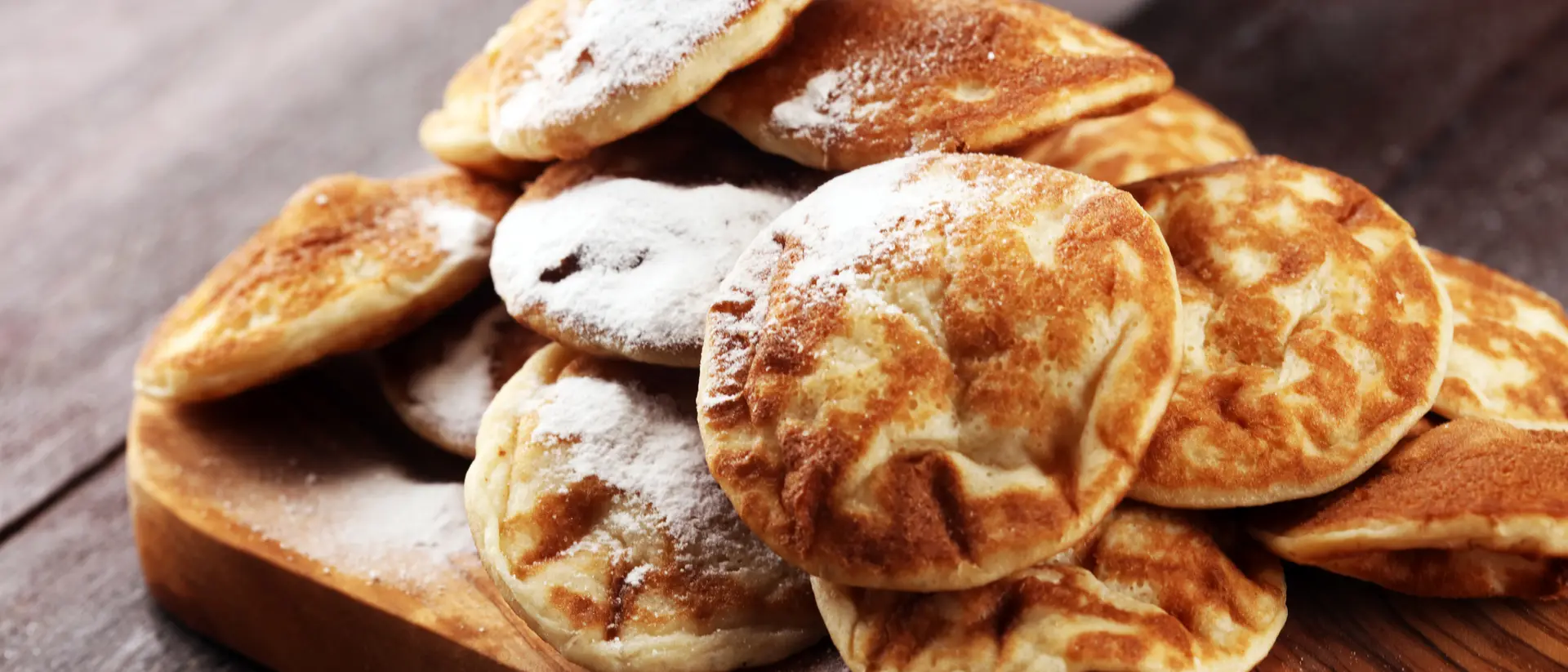 A pile of fluffy, round pancakes dusted with powdered sugar on a wooden cutting board.