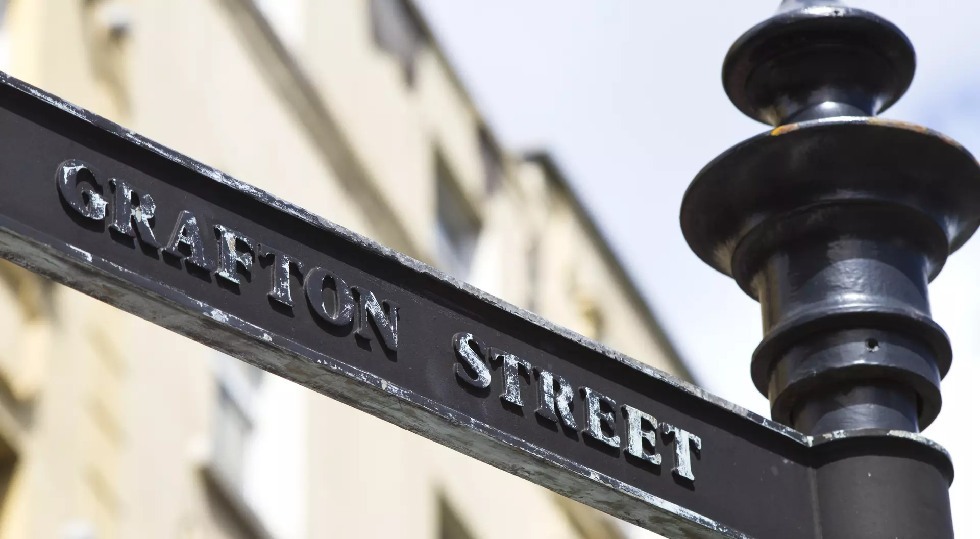 Black street sign reading "Grafton Street" against a blurred, light-colored building background.