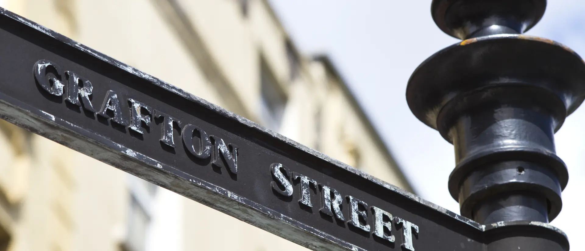 Black street sign reading "Grafton Street" against a blurred, light-colored building background.