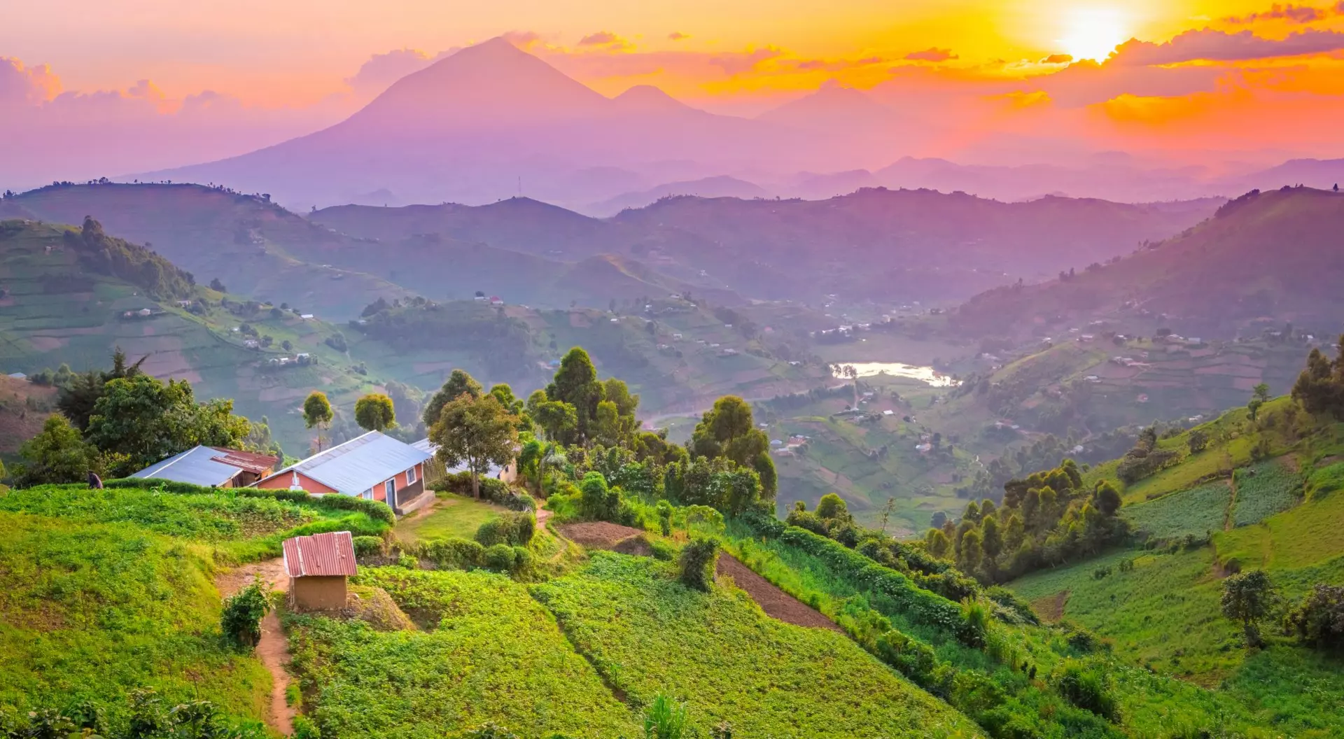Sunrise over rolling hills with lush greenery and a distant mountain, featuring a small cluster of houses.