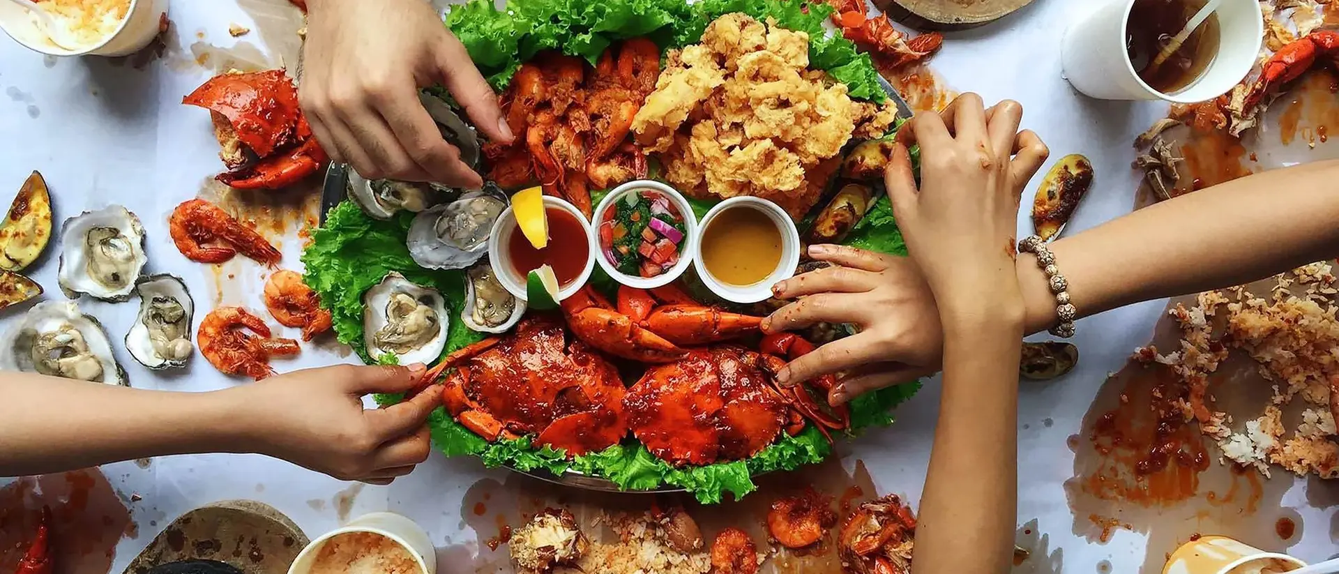 A feast of seafood, including lobsters and oysters, with hands reaching for food on a messy table covered in shells and sauces.