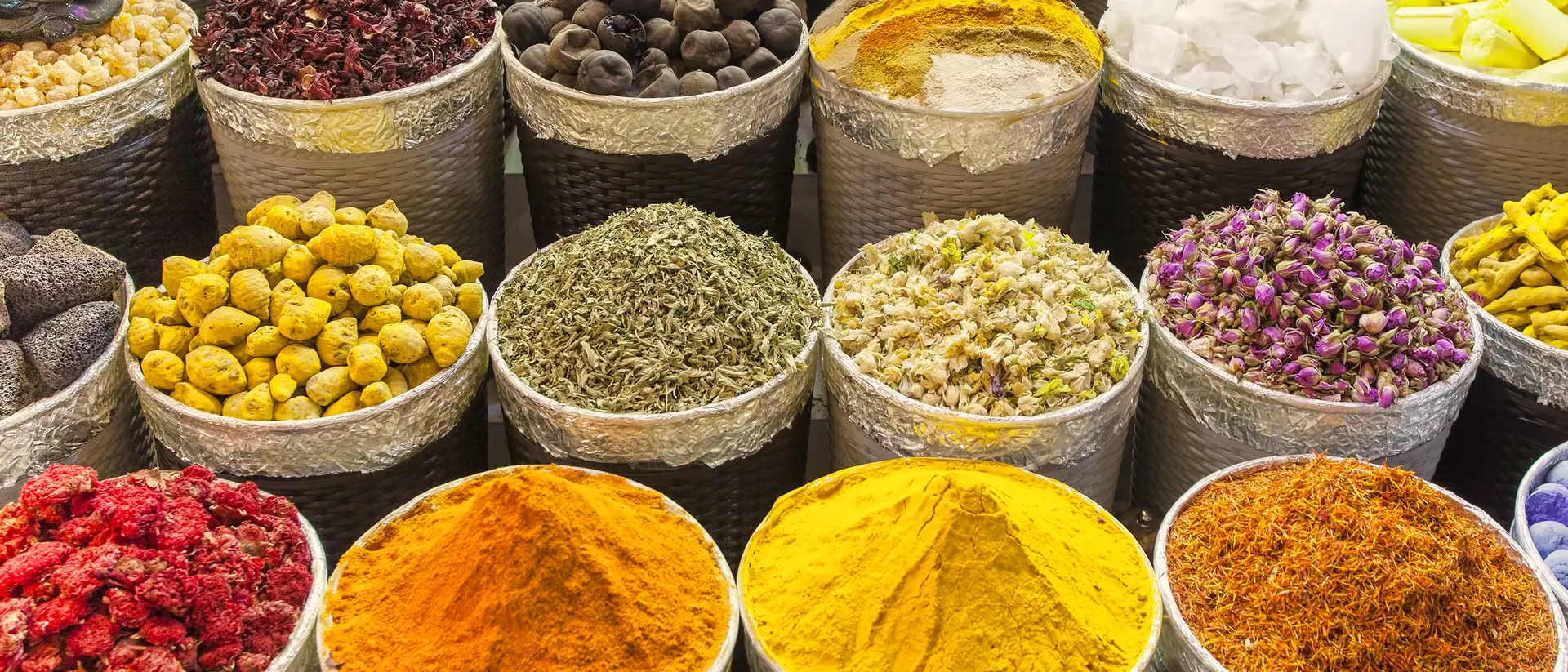 Colorful display of spices in metal containers, including saffron, turmeric, dried herbs, and floral petals.