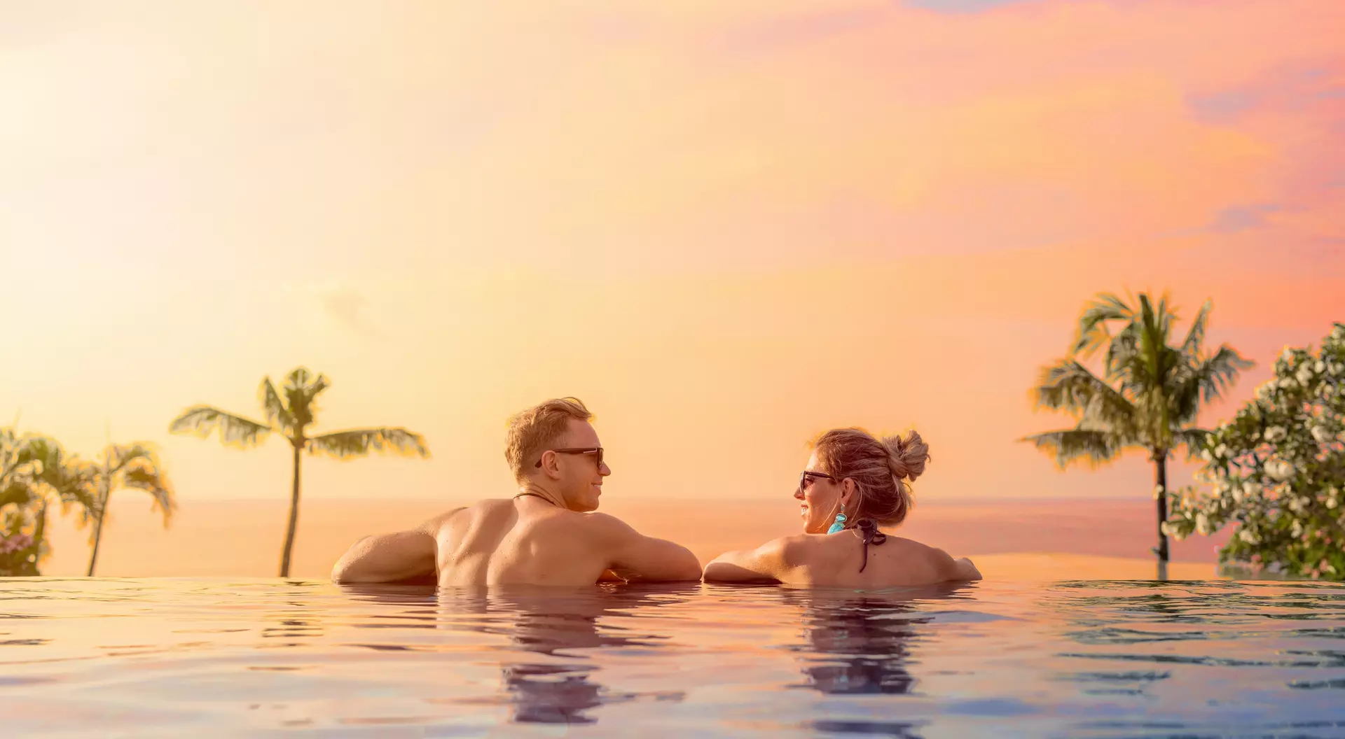A couple relaxes in an infinity pool, watching a colorful sunset with palm trees in the background.