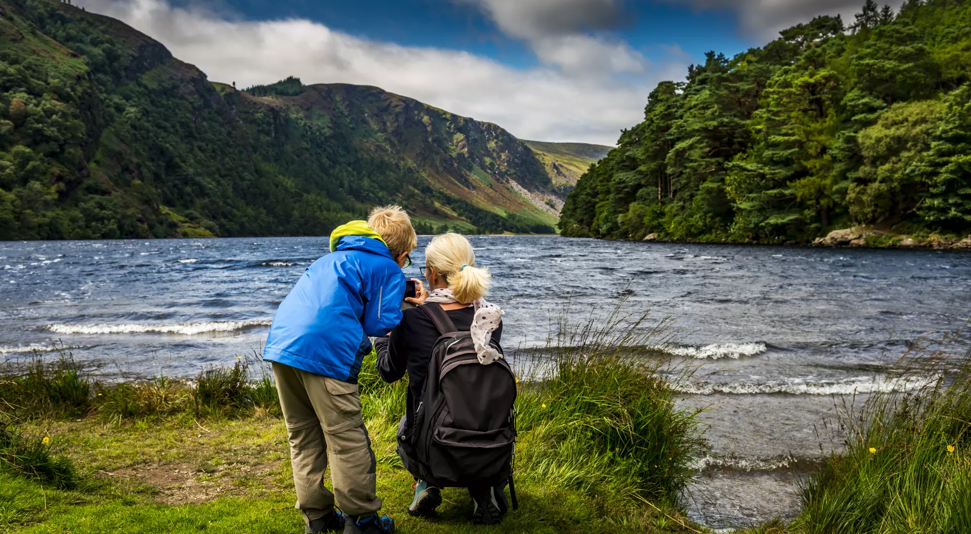 Two children, one in a blue jacket, explore a lush riverside landscape with mountains in the background.