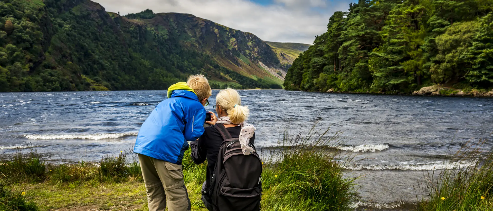 Two children, one in a blue jacket, explore a lush riverside landscape with mountains in the background.