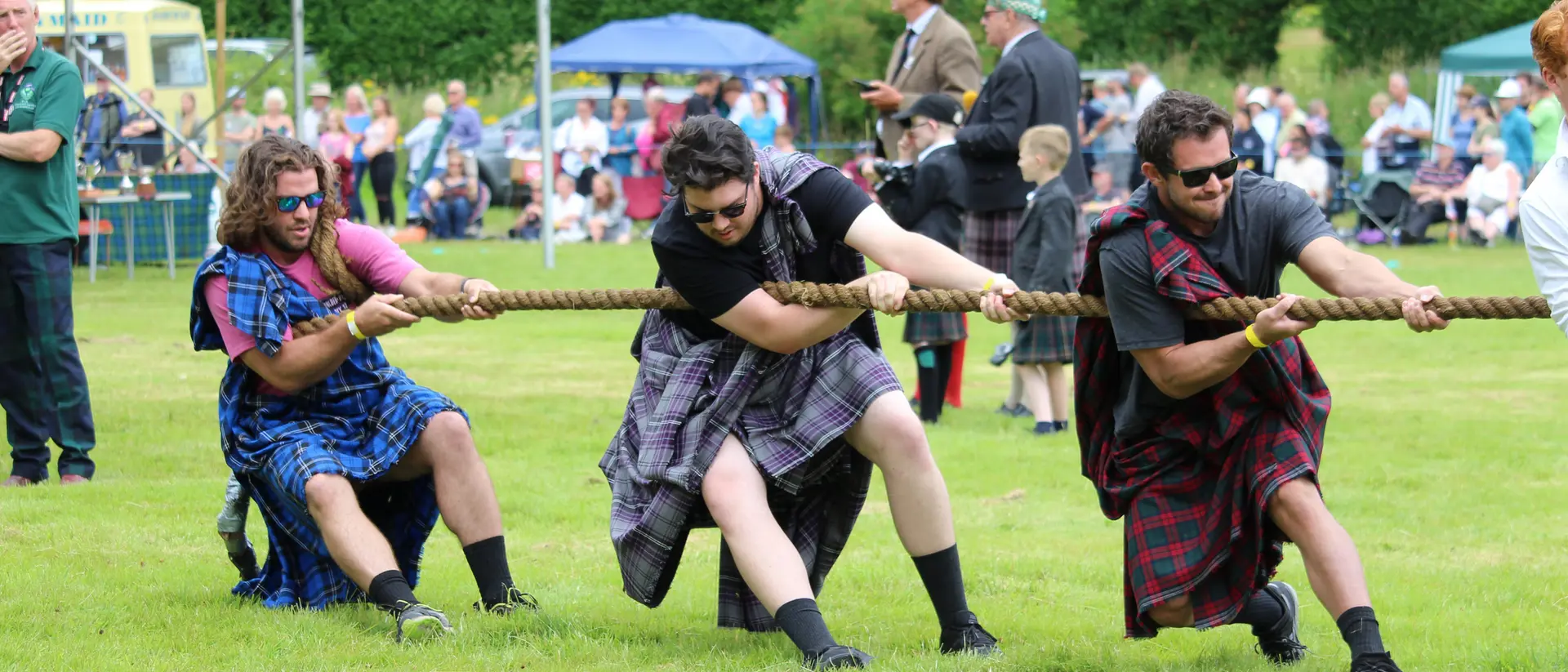 Three men in kilts are pulling a rope in a tug-of-war competition, with a crowd watching in the background.