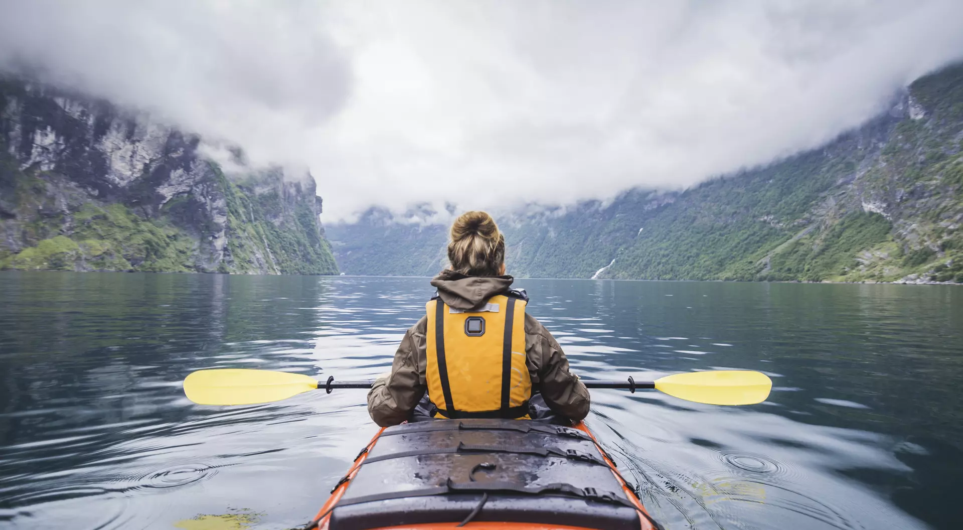 Person in a kayak on calm water, surrounded by steep cliffs and gray, cloudy skies.
