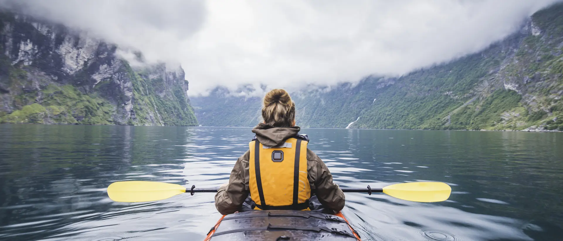 Person in a kayak on calm water, surrounded by steep cliffs and gray, cloudy skies.