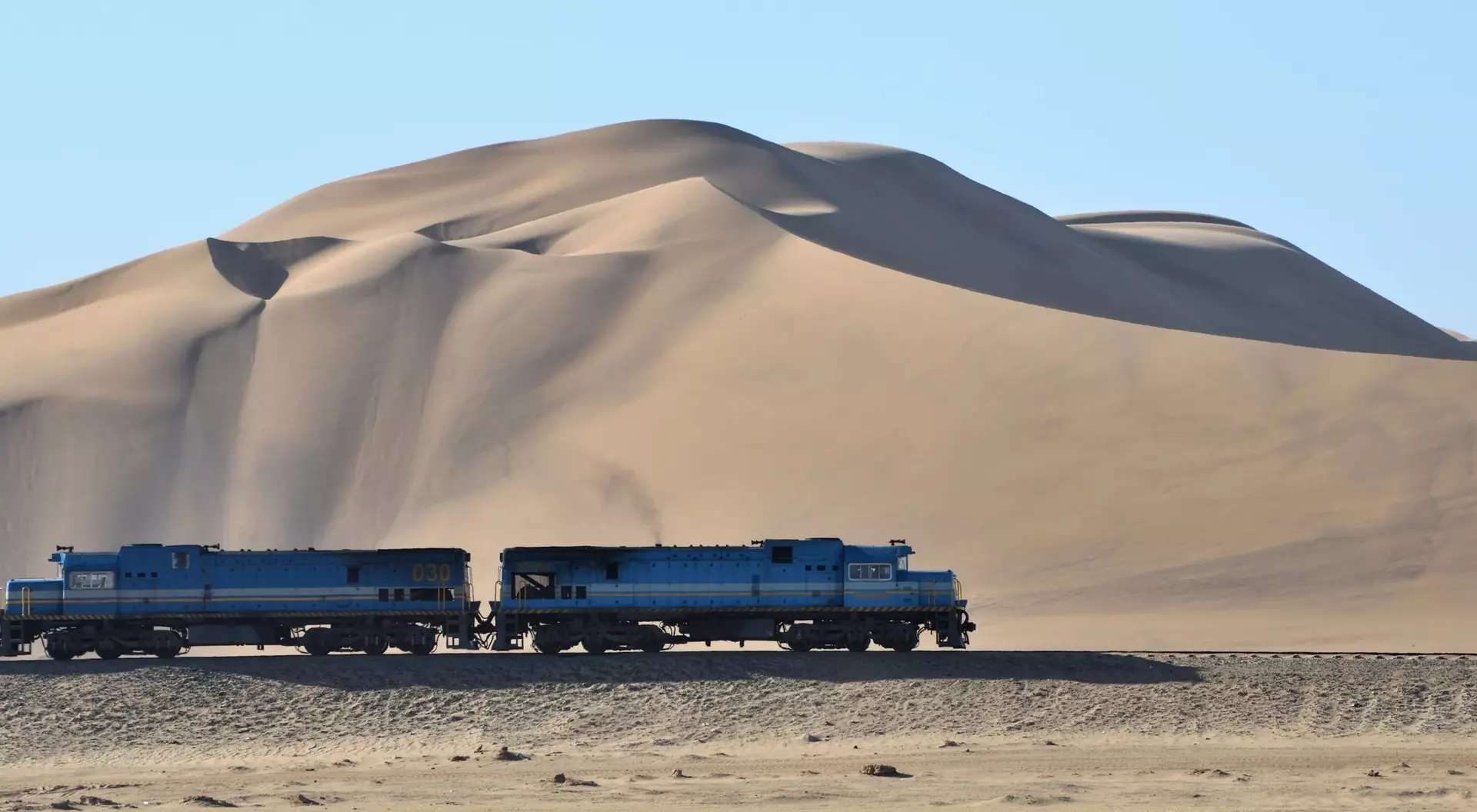 Two blue trains pass by a large sand dune under a clear blue sky.