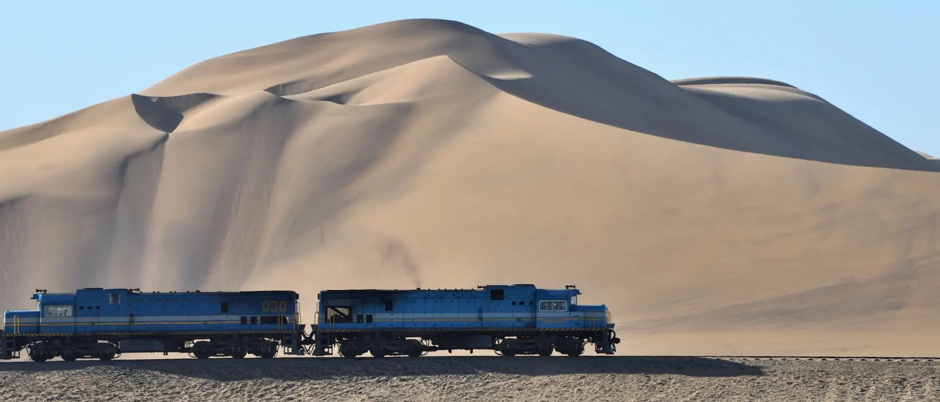 Two blue trains pass by a large sand dune under a clear blue sky.