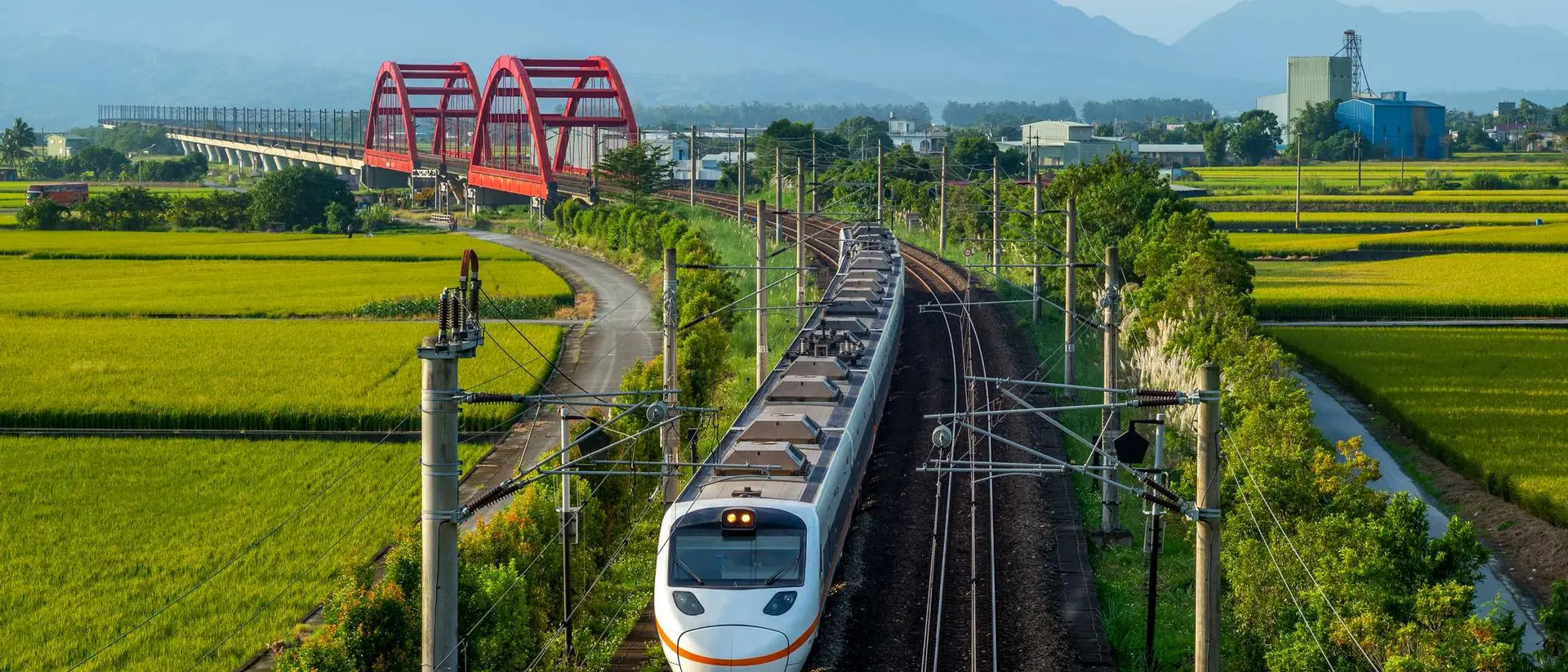 A train travels through a green landscape with rice fields and a red bridge in the background, under a clear blue sky.