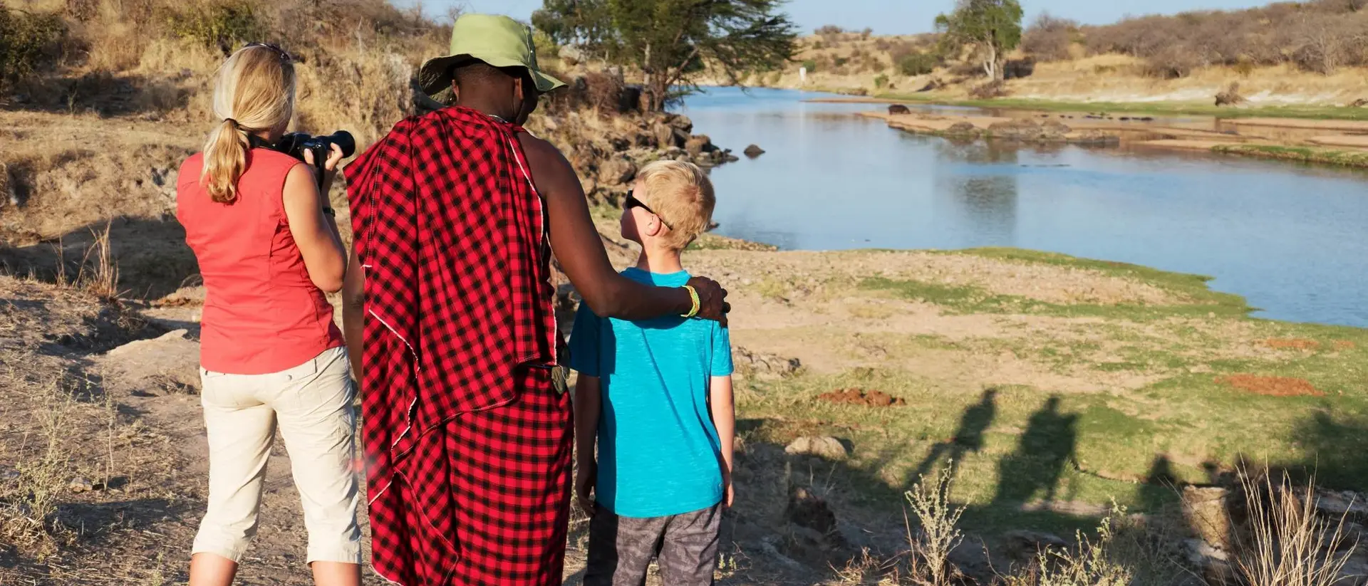 A woman and a boy watch a river with a man in traditional attire, capturing the moment as the sun sets.