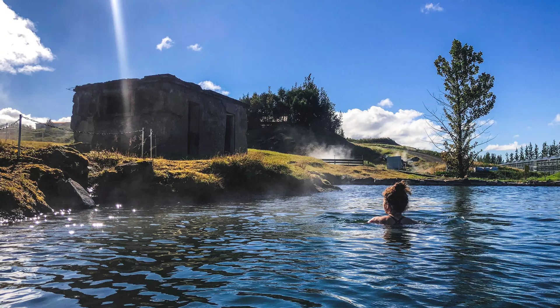 Person swimming in a natural hot spring, with a rustic building and trees under a bright blue sky.