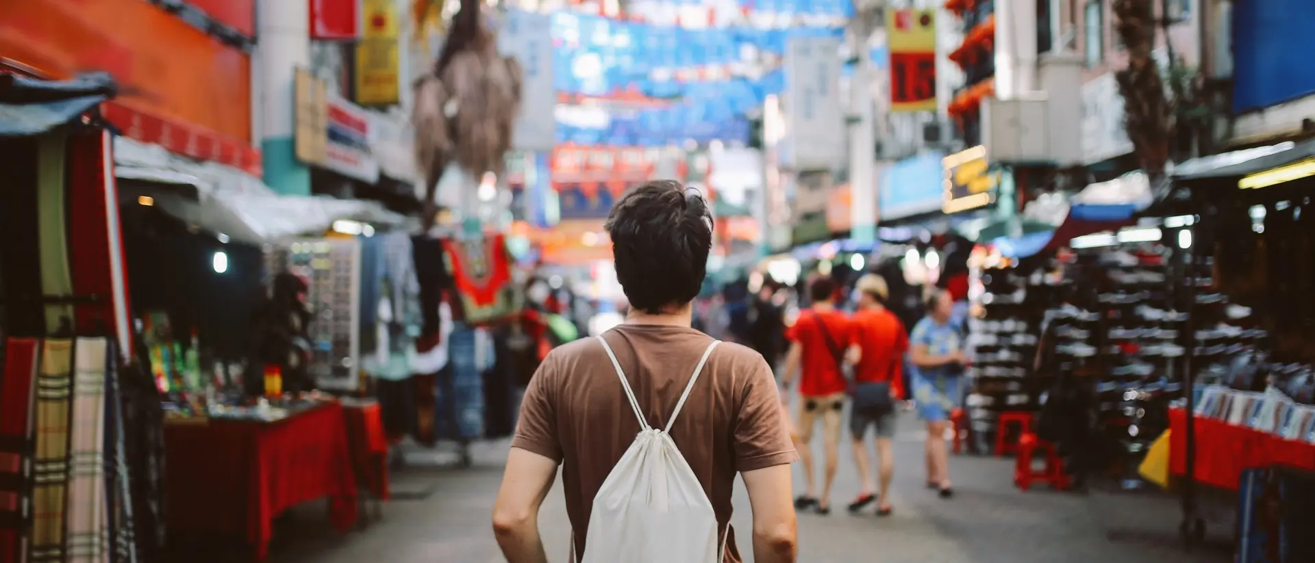 A person stands in a bustling market, facing colorful stalls and decorations, with many people in the background.
