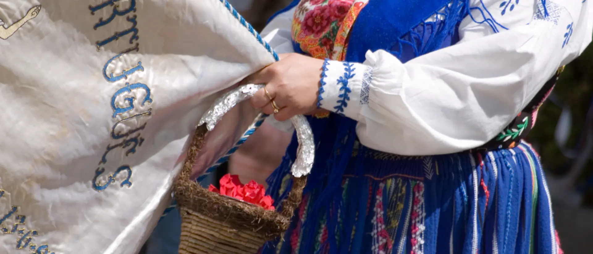 A person in traditional clothing holds a banner and a small basket of flowers during a cultural event.