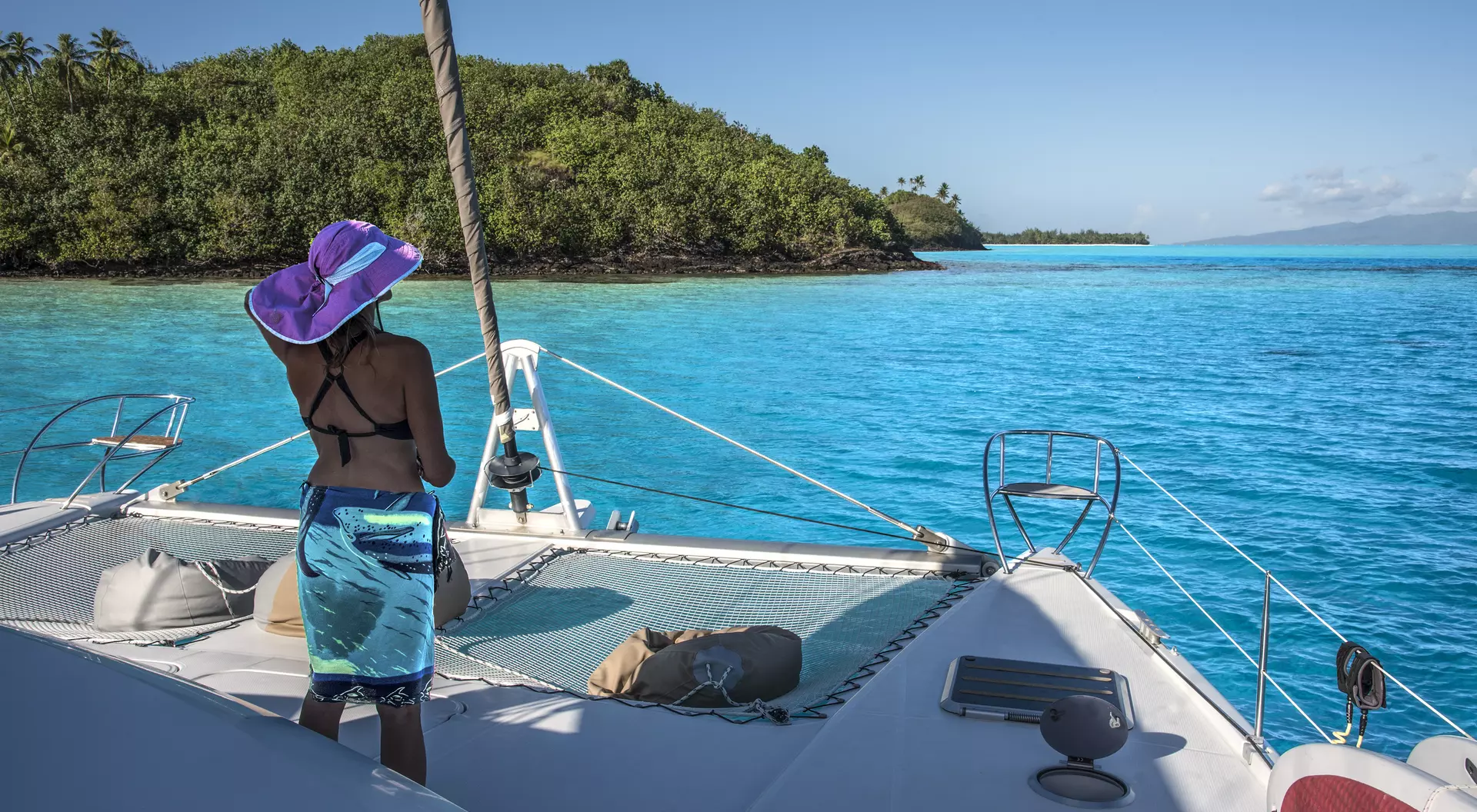 A person in a purple hat stands on a catamaran, overlooking turquoise waters and green islands under a clear sky.