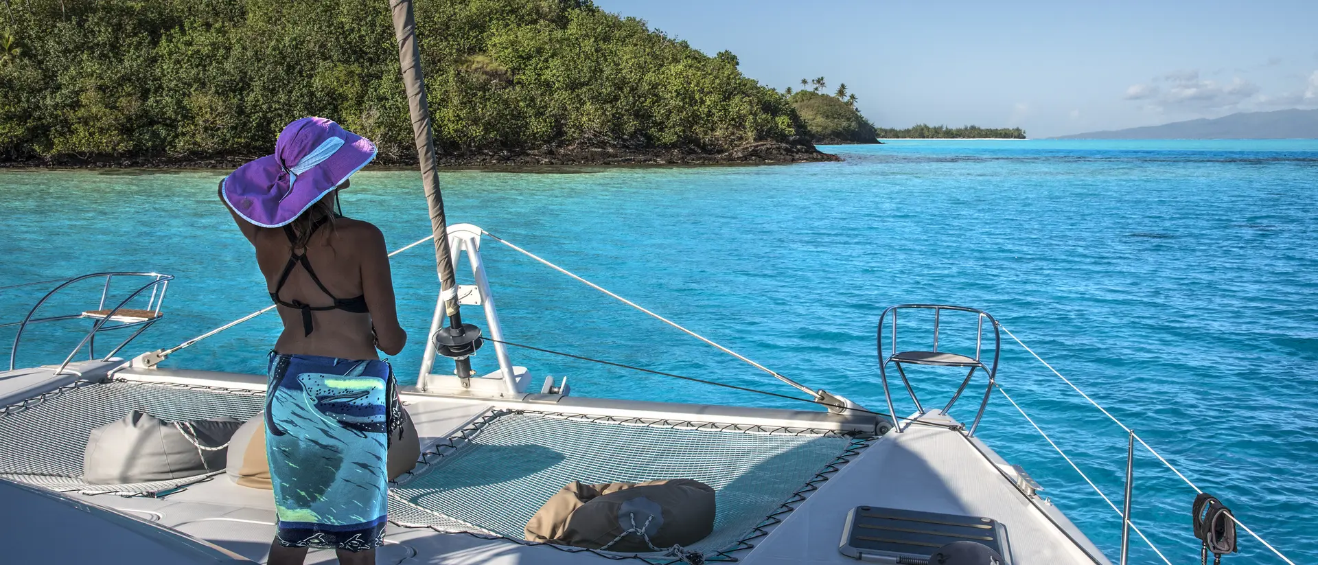 A person in a purple hat stands on a catamaran, overlooking turquoise waters and green islands under a clear sky.