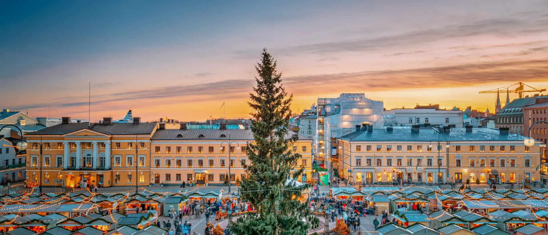 A vibrant market square at sunset, featuring a large Christmas tree and festive stalls filled with visitors and decorations.