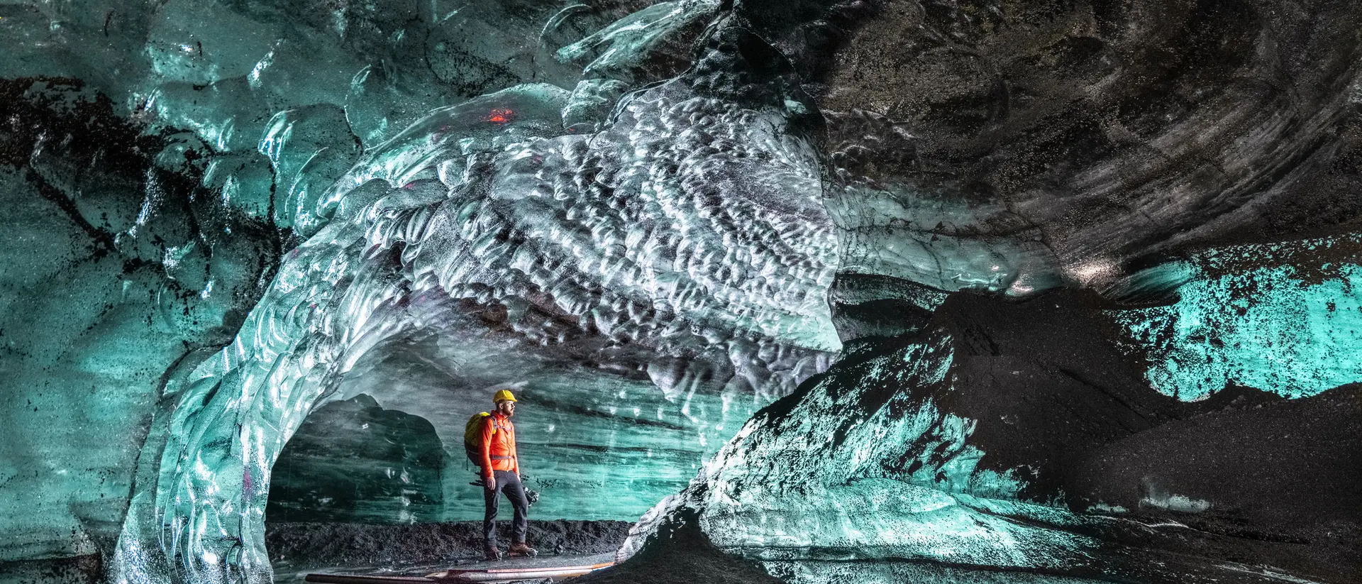A person in an orange jacket stands inside a stunning ice cave with textured blue ice formations and intricate patterns.