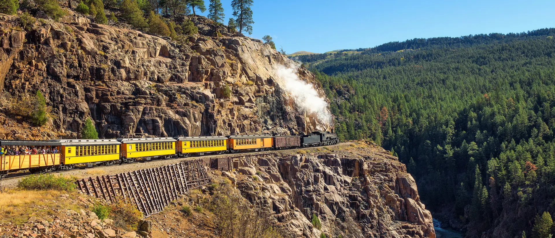 A steam train travels along a rocky cliffside, surrounded by lush green trees and a river below.