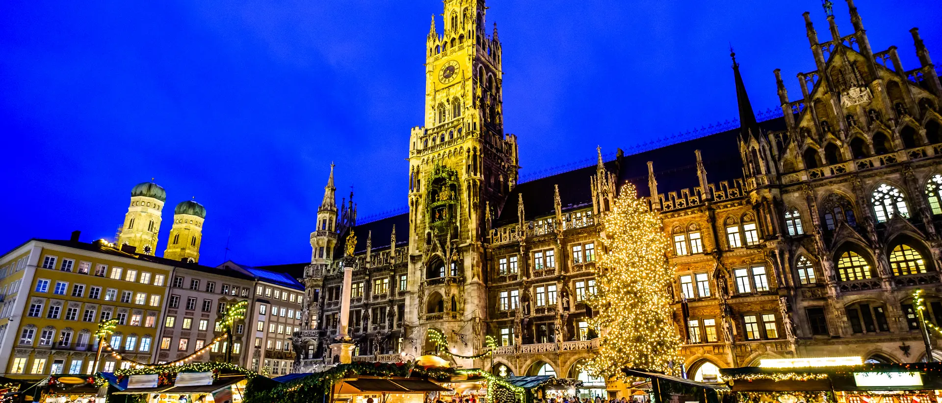A bustling market scene at night with a tall clock tower and a lit Christmas tree in the background.