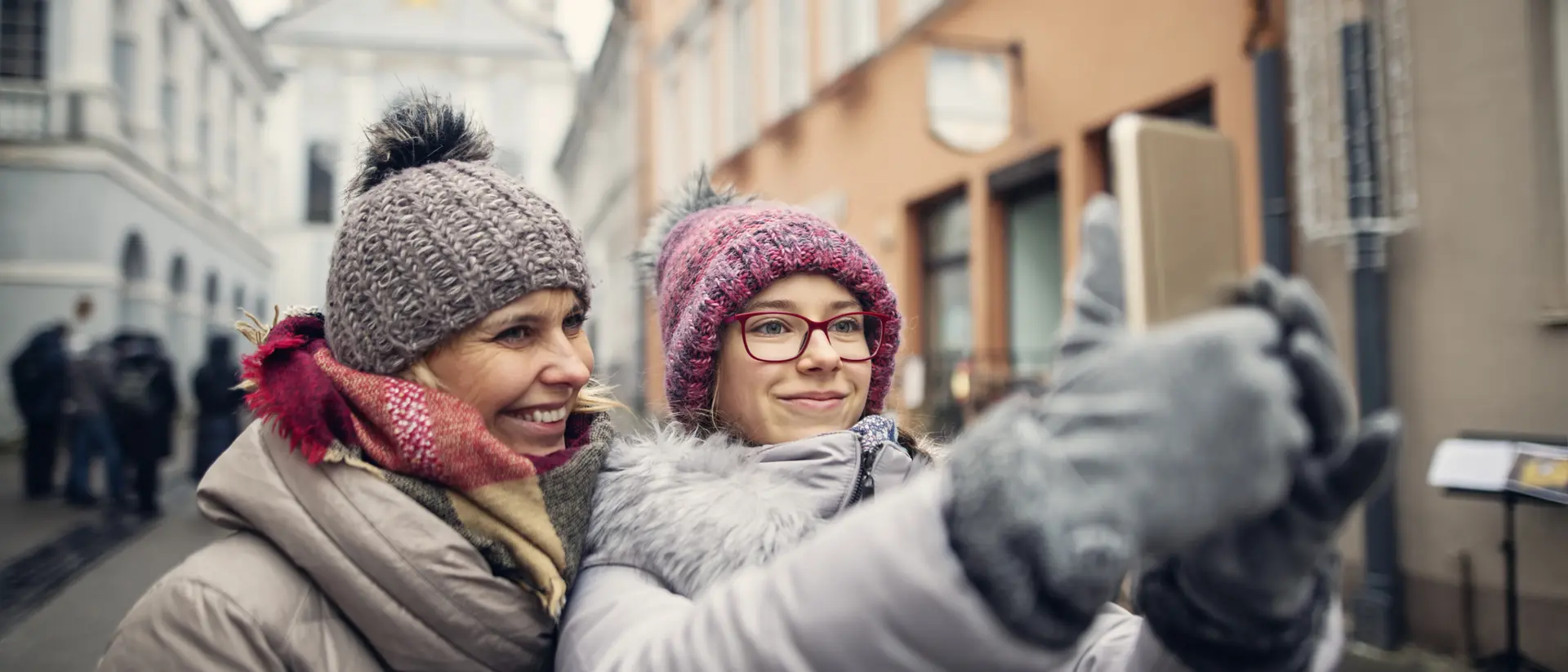 Two women in winter attire taking a selfie on a city street with historic buildings in the background.