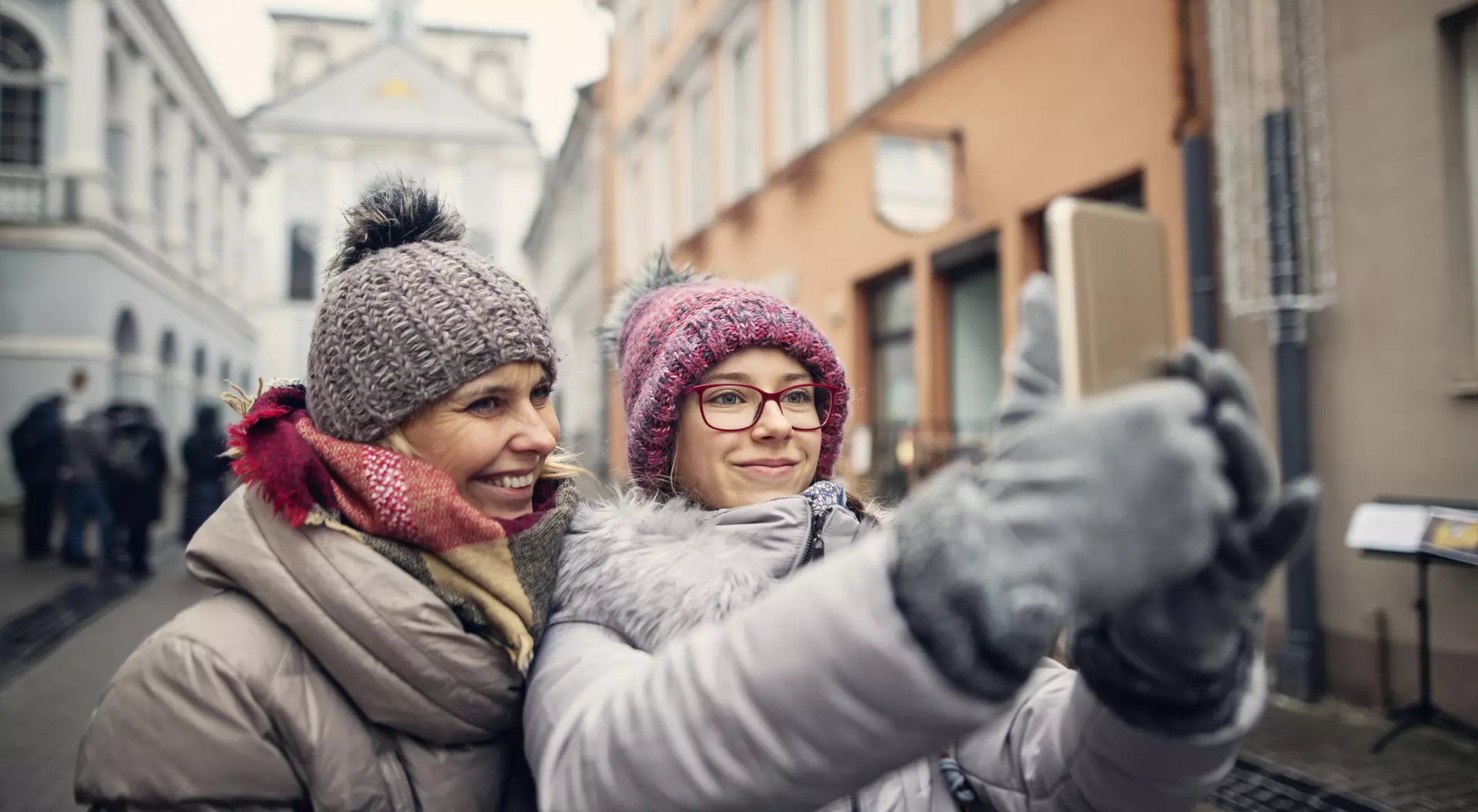 Two women in winter attire taking a selfie on a city street with historic buildings in the background.